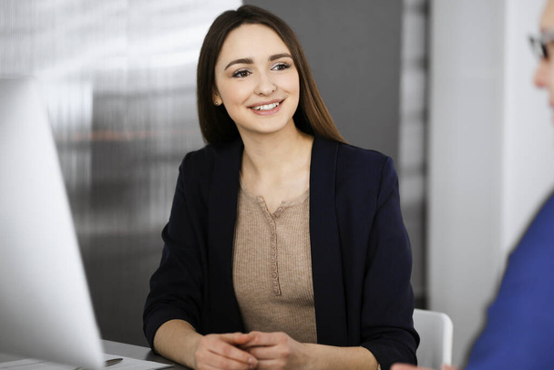 Young beautiful business woman is talking to her colleague, while sitting at the desk in a modern office. Concept of business success - Photo, Image
