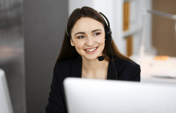 Young friendly girl in headsets is talking to a firms client, while sitting at the desk in a modern office together with her colleague. Call center operators at work - Фото, изображение