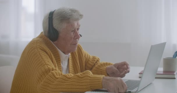 Senior žena s notebookem volá na smartphone doma. Stará žena má videohovor na notebooku, usmívá se a šťastně si povídá v útulném bytě. lékařské video volá starší pacient - Záběry, video