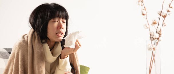 Woman wrapped in a plaid looks sick, ill, sneezing and coughing sitting at home indoors - Photo, Image