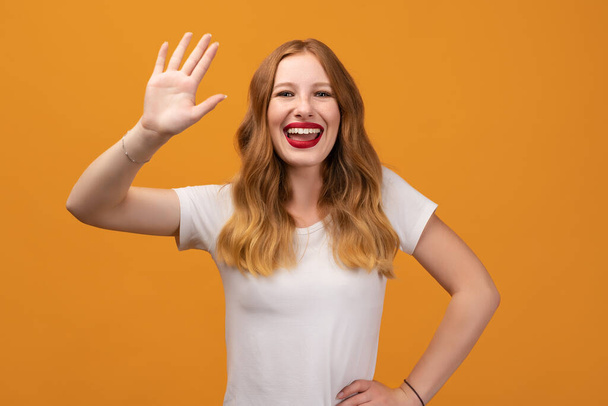 ¿Hola? Retrato de una adorable mujer amigable con pelirroja ondulada, vistiendo una camiseta blanca, de pie saludando con la mano, mirando a la cámara con una sonrisa dentada atractiva - Foto, imagen