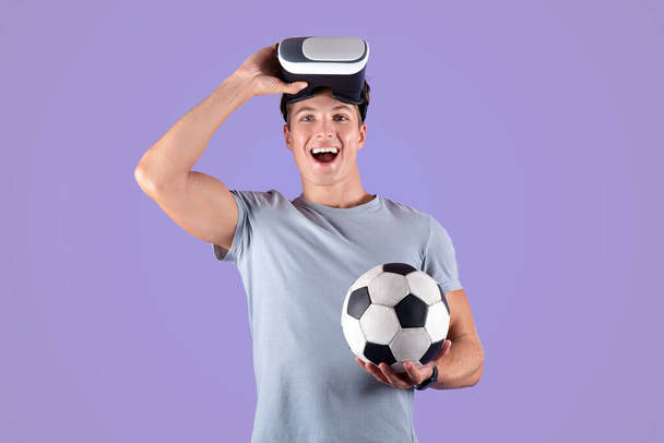 Ćwiczenia z koncepcją rzeczywistości wirtualnej. młody atletyczny facet w VR zestaw słuchawkowy trzymając piłkę nożną na liliowym tle studio - Zdjęcie, obraz