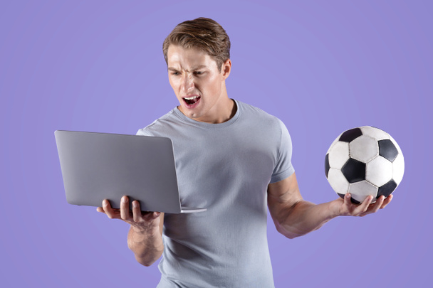 Joven decepcionado por su derrota en el equipo de fútbol, viendo el partido de fútbol en el ordenador portátil, fondo de estudio lila - Foto, Imagen