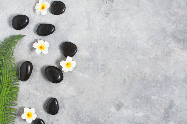 Black spa stones with Bali flowers and palm leaf top view on grey concrete background. Meditation stone background. Relaxation spa treatment concept. - Fotó, kép