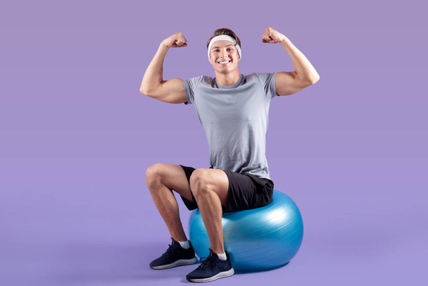 Pozytywny młody sportowiec siedzi na piłce fitness i pokazuje swoją siłę na tle studia liliowego - Zdjęcie, obraz