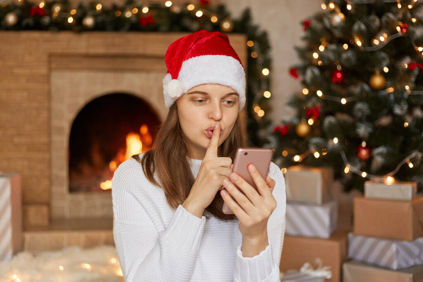 Woman in santa claus hat holding mobile phone in hands, looks at its screen, having video call, asks somebody too keep her secret, keeping finger near lips, lady wearing white sweater, posing indoor. - Foto, imagen