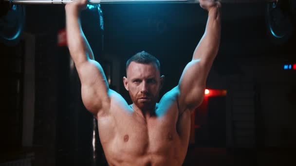 Hombre duro de pie en el gimnasio y levanta una barra pesada y bombea los músculos de sus manos sobre su cabeza - Imágenes, Vídeo