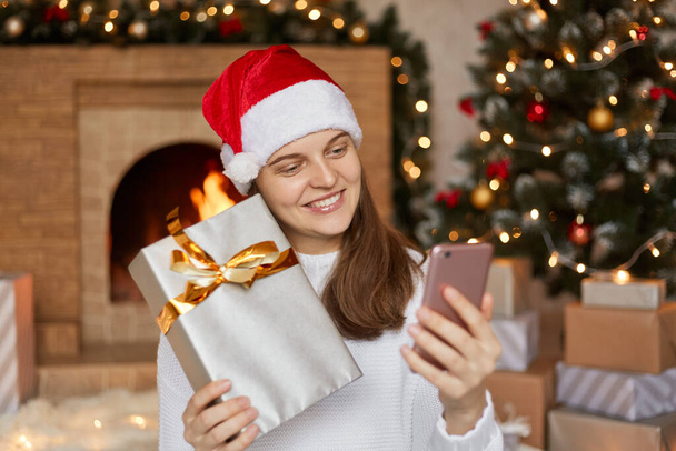 Young Caucasian woman has video call online with somebody and showing gift box in Christmas day on smart phone at home, social distancing, female video conference, communication and holiday. - Photo, Image