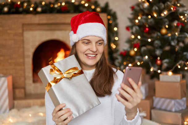 Photo of charming cute beautiful attractive girl holding her phone and gift box in hands, smiling female has video call or selfie with her Christmas present, posing in decorated living room. - Fotoğraf, Görsel