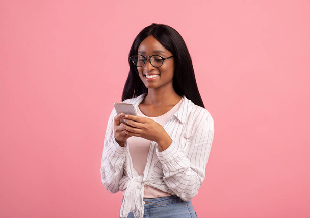 Vida online. Mulher preta inteligente em óculos usando celular para trabalho remoto ou estudos em fundo de estúdio rosa - Foto, Imagem