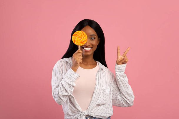 Young black woman covering her eye with bright lollipop and showing PEACE gesture on pink studio background - Foto, Bild
