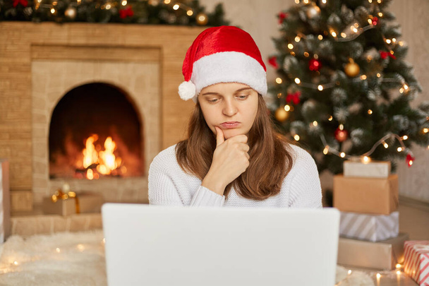 Woman in Santa hat and white sweater with laptop, has thoughtful facial expression, keeping hand on chin, looks at notebook's screen doing her freelance work during Christmas holiday. - Foto, imagen