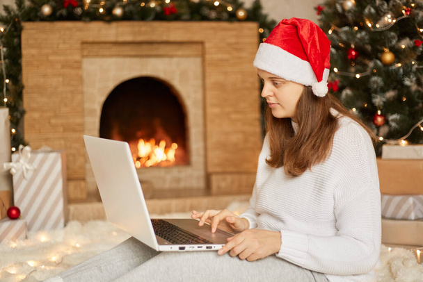 Navidad en línea, trabajando a través de Internet. Chica caucásica en casa utilizando portátil portátil portátil para videollamada, lleva sombrero de Santa y suéter blanco. Mujer mirando a la cámara del ordenador portátil, se sienta cerca de la chimenea - Foto, imagen