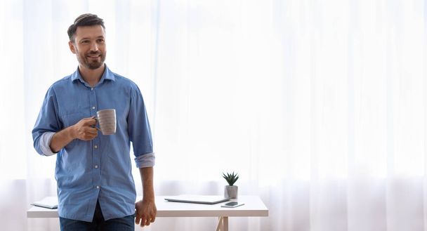 Jóképű szabadúszó srác egy csésze kávéval otthon az asztalnál. - Fotó, kép