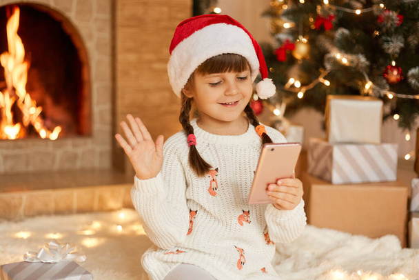Christmas online, congratulations from home, smiling little girl using smart phone for video call. Child talks to friends and parents, waves hands to greet, wearing santa hat, poses in festive room. - Zdjęcie, obraz