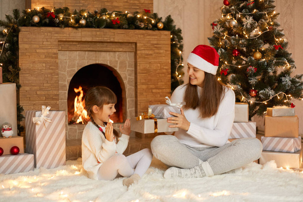 Young woman presenting Christmas gift to her little daughter at Christmas morning, fun, happy time together, excited child being very glad to get present boxes, mom in santa claus hat looks at her kid - Foto, afbeelding