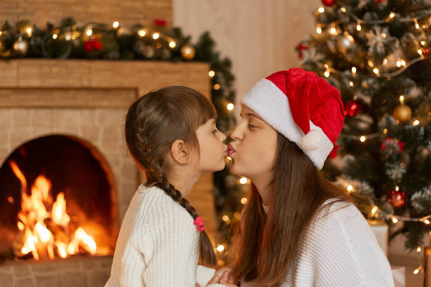 Young attractive mother and daughter posing in living room near xmas tree and fireplace, little girl kiss her mom and congratulates with holidays, wearing white jumpers. - Фото, зображення