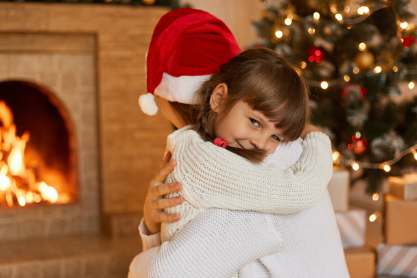 Mother and little child playing in Christmas, ladies wearing white sweaters, charming kid looks at camera with smile, kid expressing her love to mom, posing in living room with fireplace and fir tree. - Фото, изображение