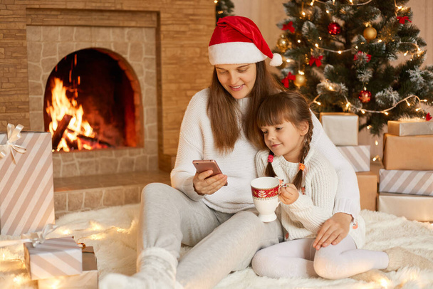 Happy mother and little daughter with smart phone at home sitting on floor, looking at device screen, having video call, posing in living room with Christmas decoration. - 写真・画像
