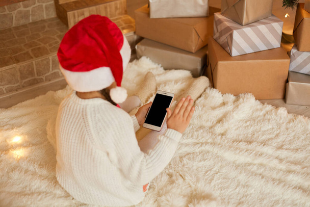 Little girl holding phone with blank screen while posing backwards, wearing white sweater and santa hat, sitting on floor on soft carpet. Merry Christmas and Happy Holidays! - Photo, Image