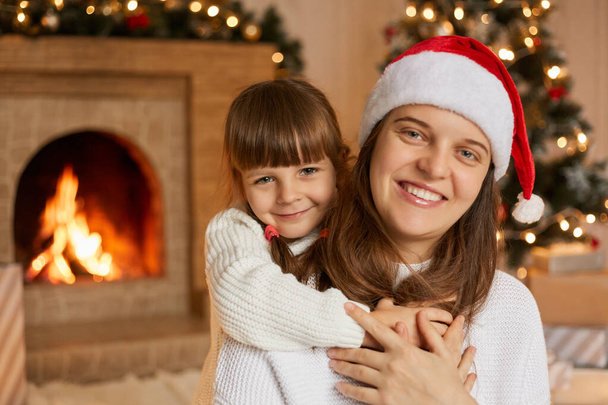 Happy family mother and little child playing in Christmas Eve, pose in living room, hugging and having fun, looking at camera with satisfied facial expressions, woman wearing santa hat. - Φωτογραφία, εικόνα