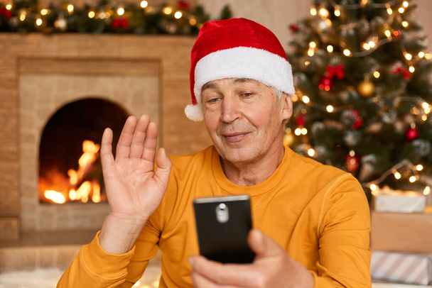 Man celebrating Christmas at home due to corona virus epidemic, holding smart phone and greeting somebody, waving hand, wearing yellow shirt and red festive hat, posing in living room with decoration. - Photo, image