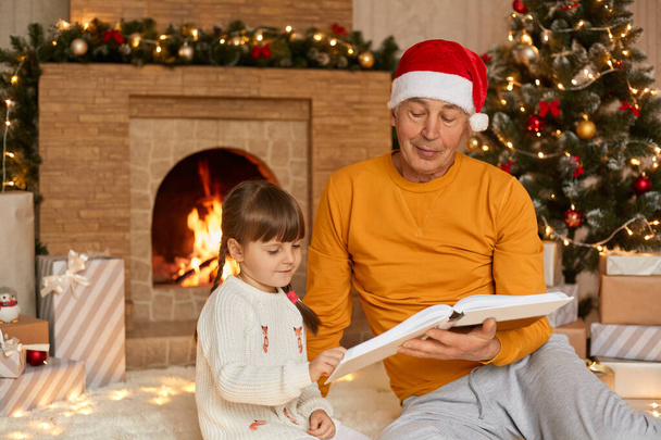 Elderly man reading book with his granddaughter in living room decorated for Christmas, family sitting on floor near fireplace and xmas tree, cute child with pigtails like spend time with grandad. - Fotó, kép