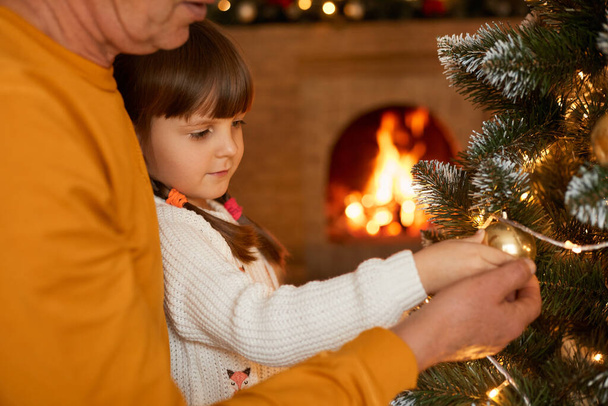 Unknown man with little daughter standing by xmas tree, decorating its with xmas ball, looking at garland's lights, posing with blurred fireplace on background. - Foto, imagen