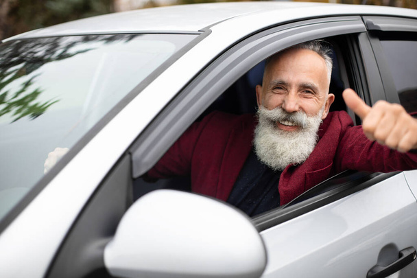 Улыбающийся пожилой бизнесмен за рулем роскошной машины, показывающий большой палец вверх - Фото, изображение