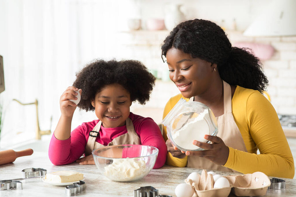 Fröhliche schwarze Mutter und kleine Tochter bereiten gemeinsam Teig in Küche zu - Foto, Bild