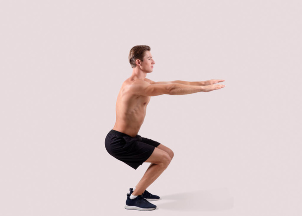 Side view of strong young guy doing squats on light studio background, full length portrait - Valokuva, kuva