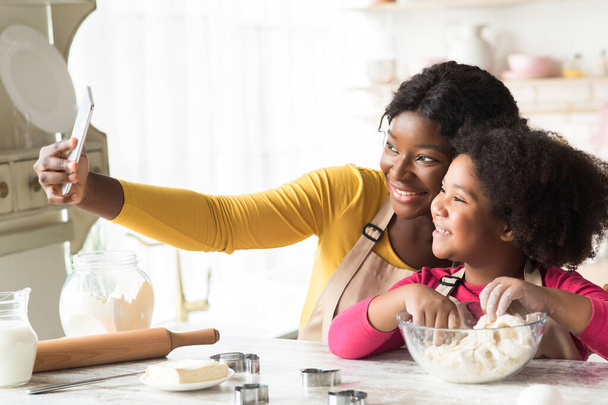 Hermosa madre negra y su hija pequeña tomando selfie en el teléfono inteligente en la cocina - Foto, imagen