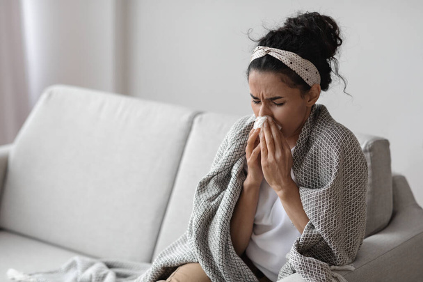 Mujer joven enferma envuelta en manta estornudando, interior del hogar - Foto, Imagen
