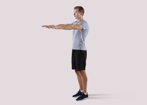 Strong millennial guy doing squats exercises on light studio background, full length portrait - Photo, Image