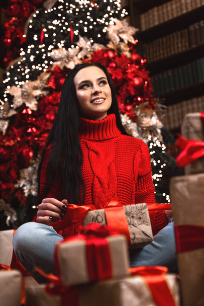 Full length photo of positive girl, hat hold big giftbox get on christmas night enjoy newyear tradition sit on floor carpet in house. - Foto, Imagen