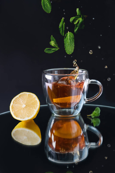 a glass cup with black tea with a splash and falling mint leaves on a black background - Foto, afbeelding