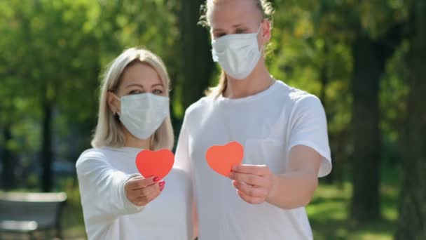 Een man met een meisje in witte T-shirts en beschermende maskers op hun gezicht tillen rode harten in het frame met hun handen, verbinden harten en kijken elkaar aan. Veilige liefde. - Video