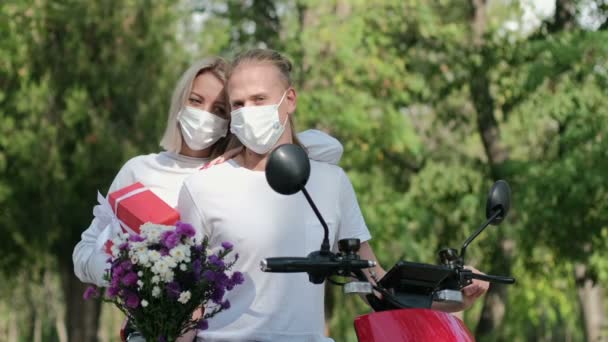 Yüzlerinde koruyucu maskeli bir kız olan bir adam parkta motosikletin yanında oturur ve maskenin içinden bir buket çiçek koklar. Gençler için güvenli bir randevu.. - Video, Çekim