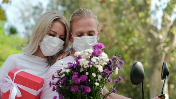 De cerca, un chico y una chica con máscaras médicas tratan de oler y saborear el olor de las flores mientras están sentados en una moto en el parque. - Metraje, vídeo