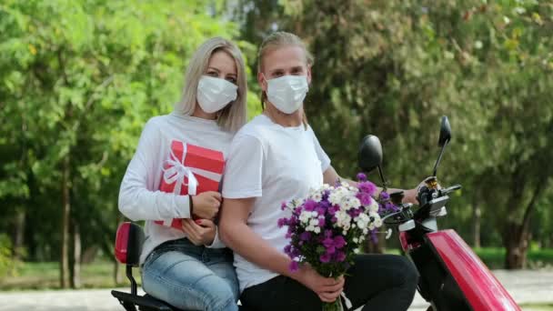 Bir adam ve koruyucu maskeli bir kız bir motosiklete binip kameraya bakıyorlar. Kız beyaz kurdeleli kırmızı bir kutuya sarılıyor ve adam elinde bir buket çiçek tutuyor.. - Video, Çekim