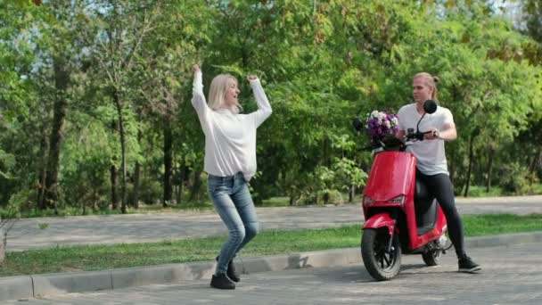 Das Mädchen tanzt im Park, der Kerl sitzt auf einem roten Moped und wirft dem Mädchen einen Blumenstrauß zu. Das Mädchen fängt einen Blumenstrauß, überrascht davon und tanzt fröhlich. - Filmmaterial, Video