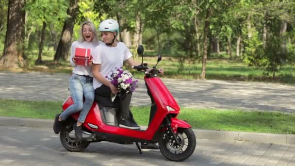 Een jong meisje met een bril knuffelt een rode doos met een wit lint en kust een man met een boeket bloemen zittend op een rode brommer in het park. - Video