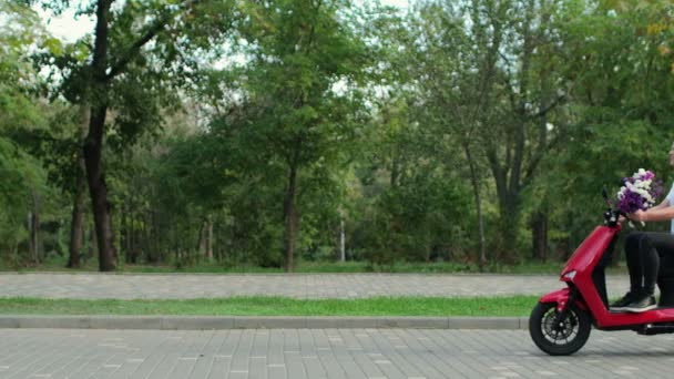 Um jovem com um buquê de flores cavalga ao longo da estrada no parque em um ciclomotor elétrico. - Filmagem, Vídeo