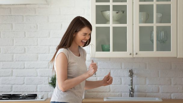 Overjoyed funny young woman singing into whisk, standing in kitchen - Fotó, kép