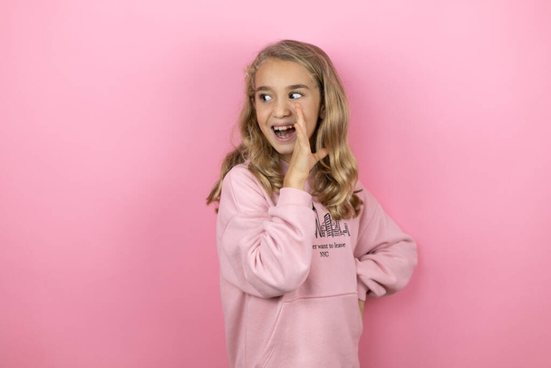 jong mooi kind meisje staande over geïsoleerde roze achtergrond met haar hand op de mond vertellen geheim gerucht, fluisteren kwaadaardig gesprek - Foto, afbeelding
