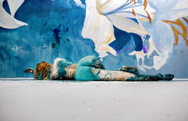 Bodypainting, back view of an artistically painted lying sexy young redheaded woman in the studio with lilies painted wall lying on the floor, copy space - Photo, Image