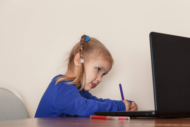 Το κοριτσάκι μελετά επιμελώς στον υπολογιστή. Το παιδί ζωγραφίζει με μαρκαδόρους. Το κοριτσάκι διαβάζει και μετά βίας βγάζει τη γλώσσα της. - Φωτογραφία, εικόνα