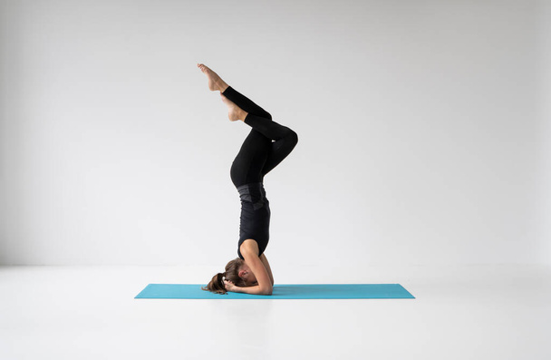 Young woman is practicing yoga in a studio. Slim young girl in a black sport wear is doing yoga practice on a blue mat. Concept of healthy life and natural balance between body and mental development. - Foto, Imagen