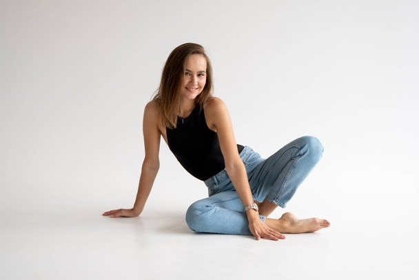 Portrait of happy young pensive woman posing in black underwear and blue jeans without a shoes, sitting on a white floor in white studio. Model tests of pretty girl in basic clothes on cyclorama. - Fotografie, Obrázek