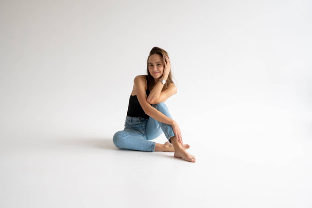 Portrait of happy young pensive woman posing in black underwear and blue jeans without a shoes, sitting on a white floor in white studio. Model tests of pretty girl in basic clothes on cyclorama. - Fotoğraf, Görsel